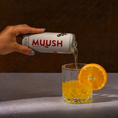 Hand pouring a can of nootropic drink MUUSH into a glass with an orange slice.