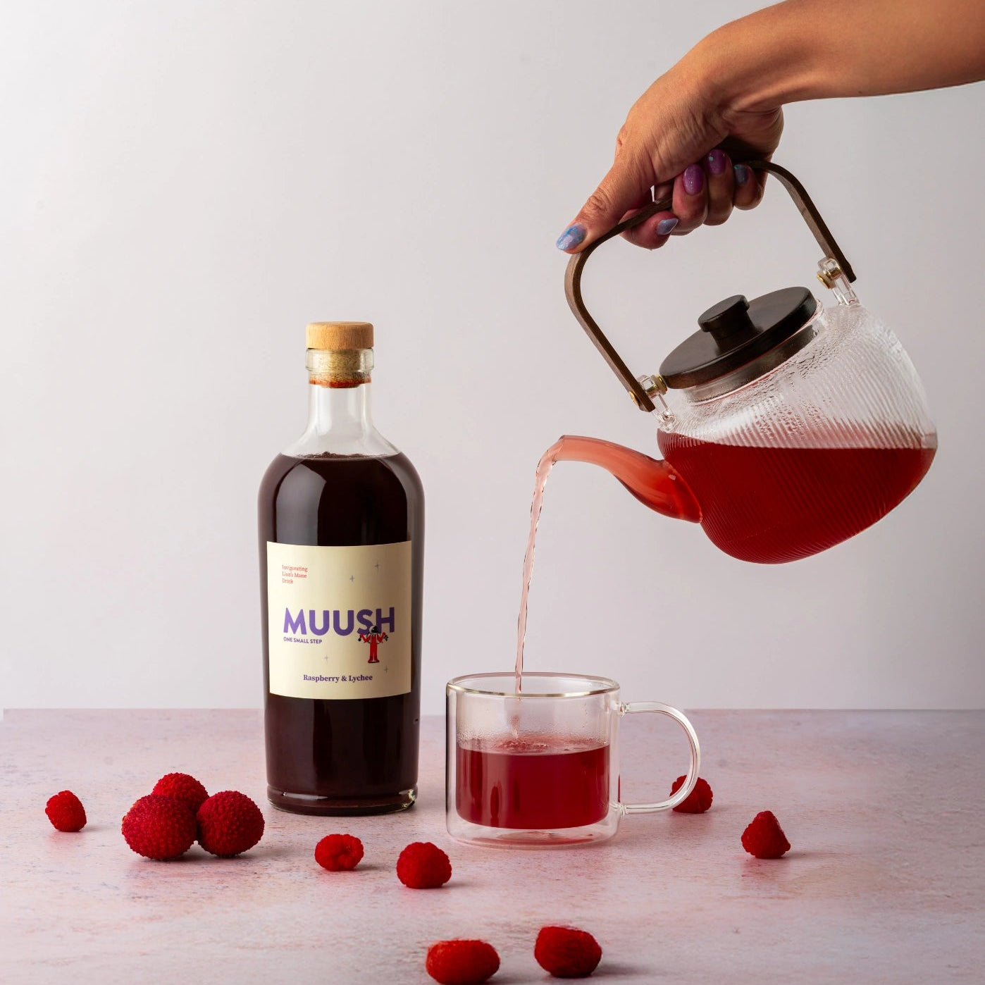 Person pouring tea made with MUUSH Raspberry and Lychee from a teapot into a glass mug next to a bottle of MUUSH Raspberry and Lychee Elixir 