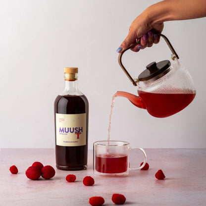 Person pouring tea made with MUUSH Raspberry and Lychee from a teapot into a glass mug next to a bottle of MUUSH Raspberry and Lychee Elixir 