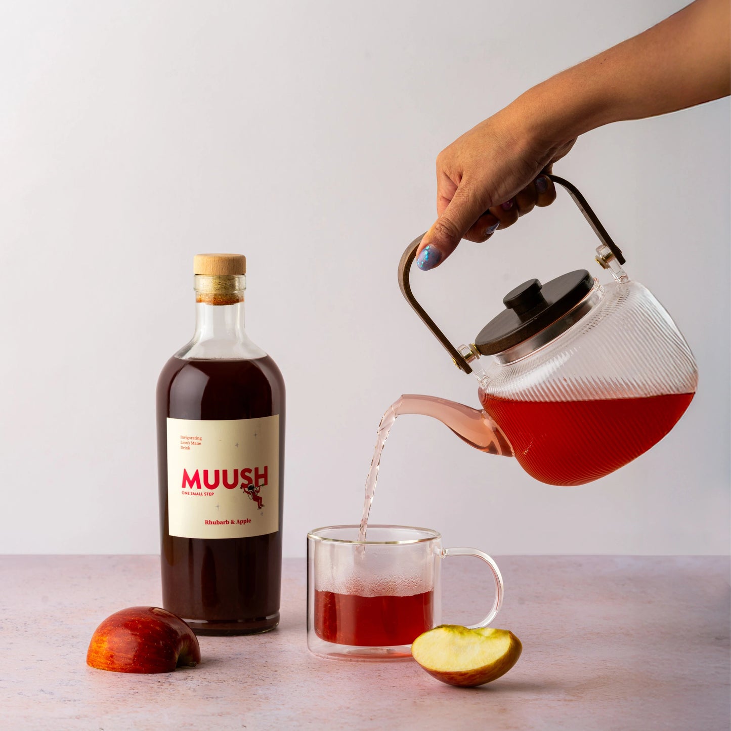 Person pouring red liquid from a teapot into a glass mug with a bottle of MUUSH rhubarb and apple on a light surface.