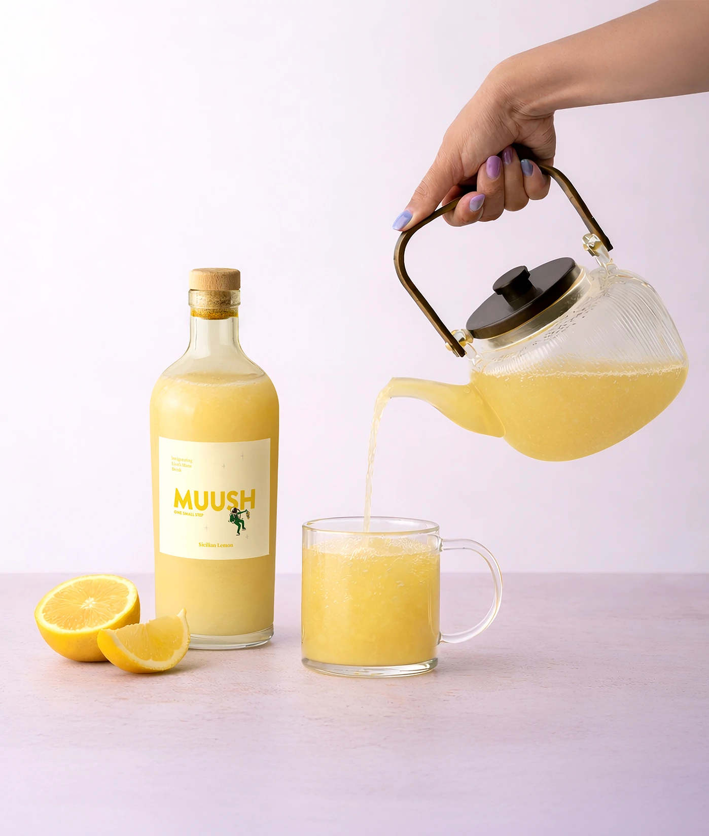 Person pouring yellow liquid from a teapot into a glass, with a bottle labeled 'MUUSH SICILIAN LEMON ELIXIR' and lemons on a light background.