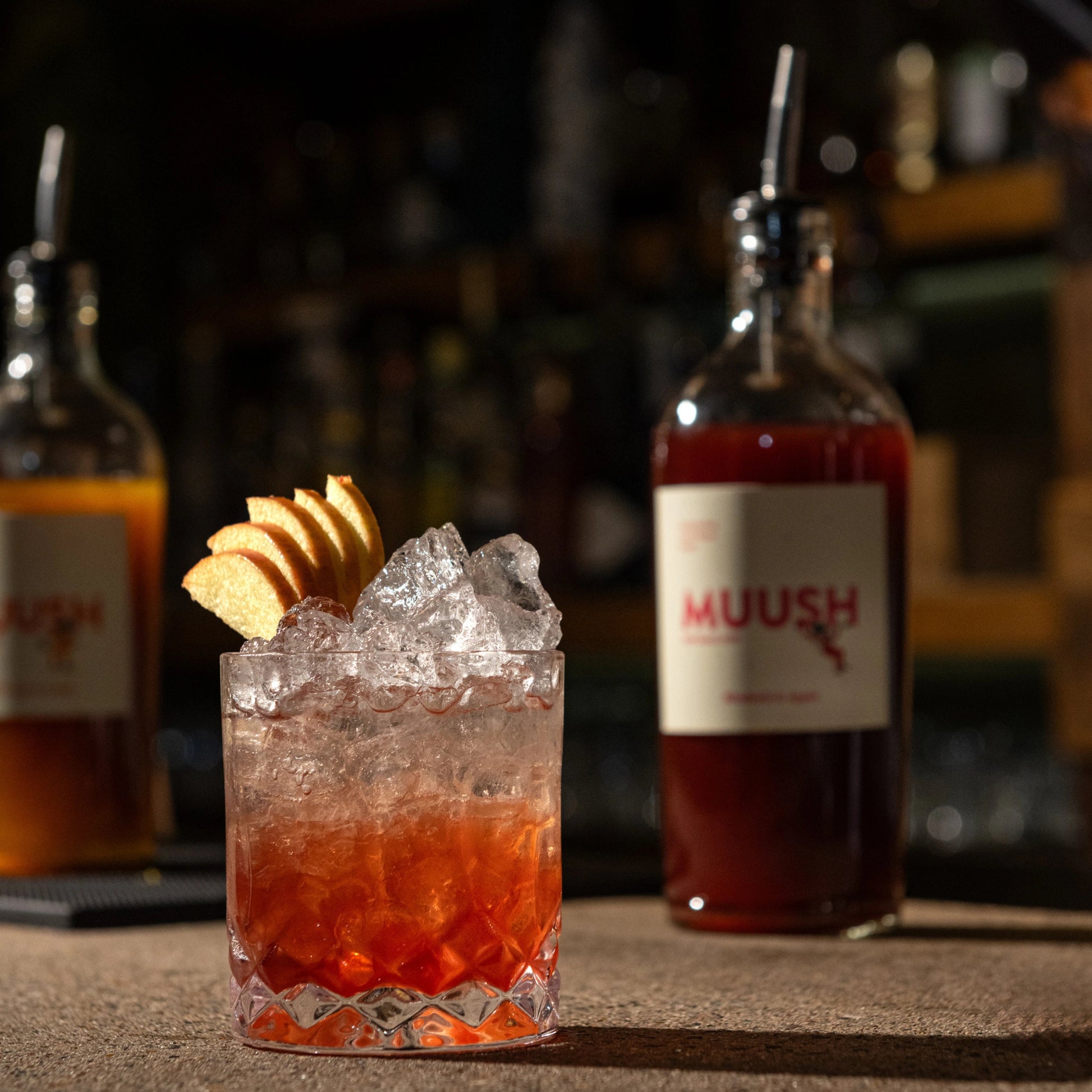 Cocktail with ice and a slice of apple garnish, Muush rhubarb and apple bottle in the background