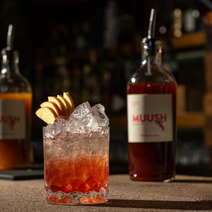 Cocktail with ice and a slice of apple garnish, Muush rhubarb and apple bottle in the background