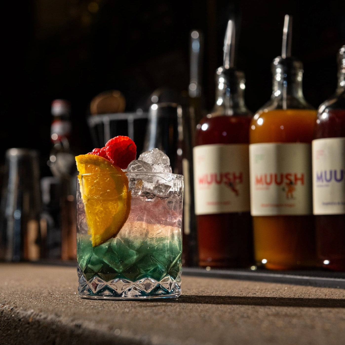 Non alcoholic cocktail with ice and fruit garnish in front of MUUSH Elixir bottles on a bar.