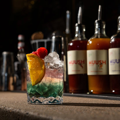 Non alcoholic cocktail with ice and fruit garnish in front of MUUSH Elixir bottles on a bar.