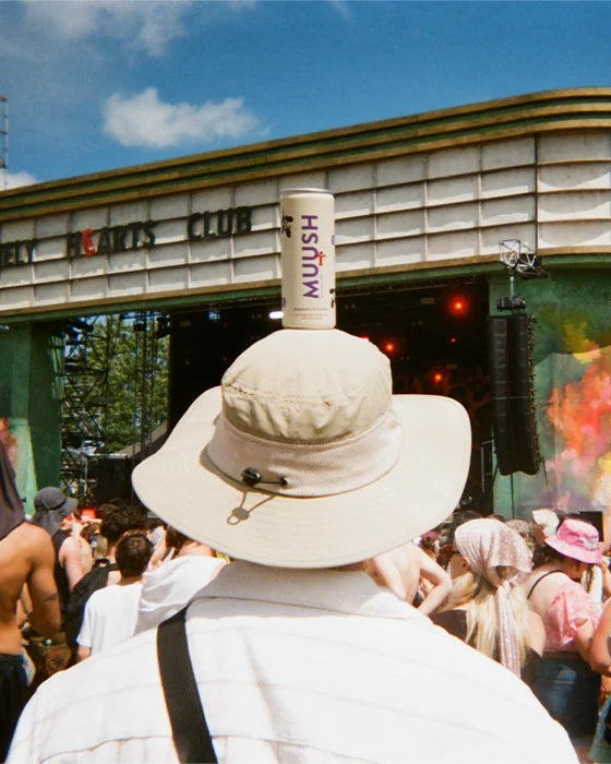 A person at an outdoor concert or club scene, seen from behind, wearing a large beige wide-brim hat with a cylindrical bottle balanced on top. The bottle has the word 'MUOSH' printed vertically.