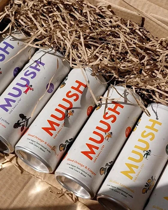 Several MUUSH sparkling tea cans arranged in brown paper shred inside a cardboard box. The visible can labels show different flavor names and colorful MUUSH branding.