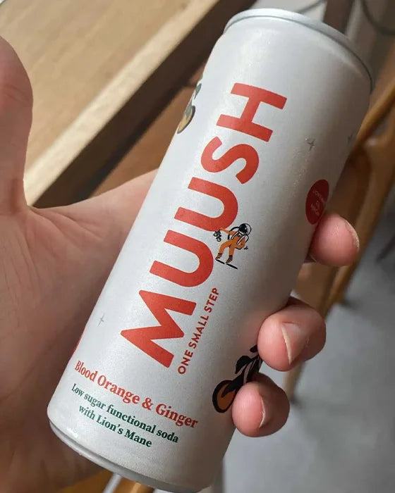 A hand holding a can of Muush Blood Orange & Ginger low sugar functional soda. The can is white with bold red lettering and a small cartoon astronaut graphic, set against a blurred indoor background.