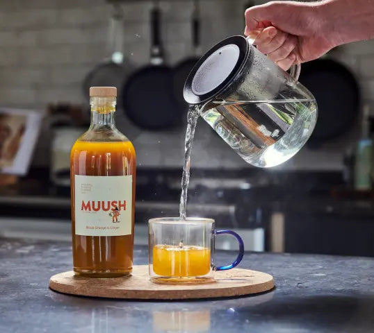 Person pouring liquid from a glass pitcher into a mug on a kitchen counter with a  'MUUSH' blood orange & ginger elixir bottle in the background
