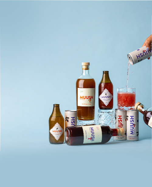 Bottles of Muush 70cl Elixirs bottles with Muush 300ml nootropic soda and Muush 250ml functional soda cans with a person pouring Muush raspberry and lychee Functional soda in a glass on a light blue background